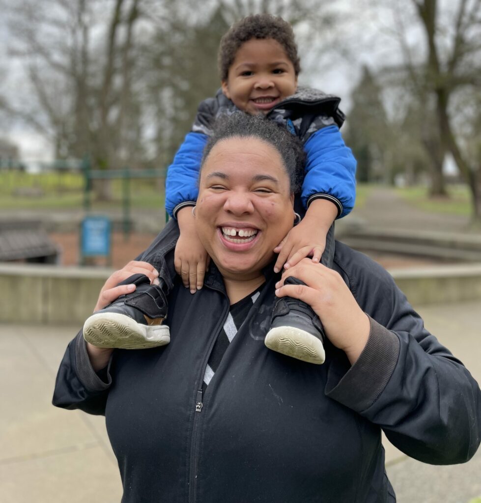Courtnee poses for a picture in a park with her son sitting on her shoulders. 